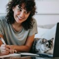 glad-woman-with-cat-writing-in-planner-while-using-laptop