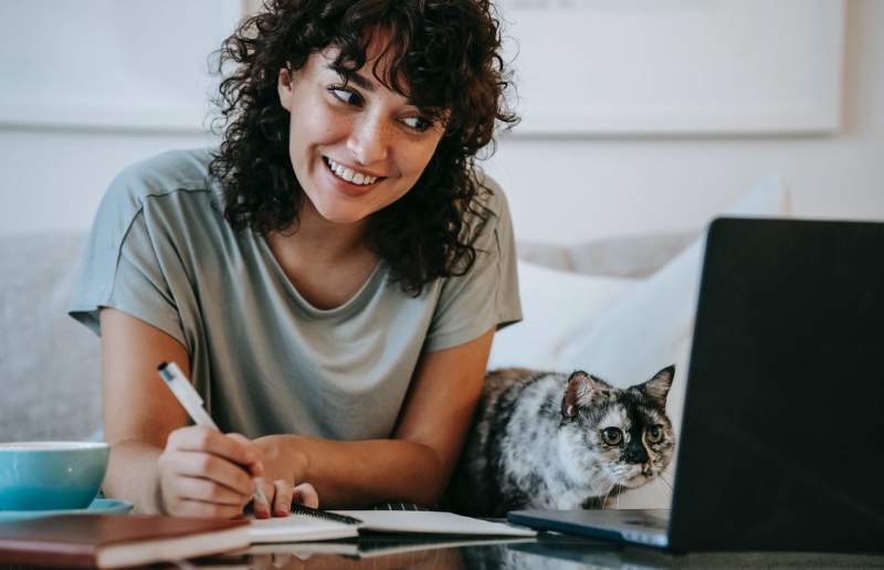 glad-woman-with-cat-writing-in-planner-while-using-laptop